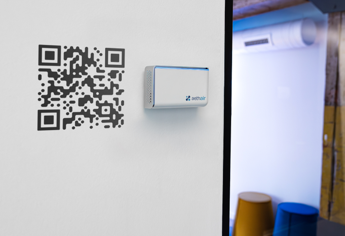 Aethair IAQ monitoring the indoor air quality of a room, mounted next to a QR code that provides real-time data access to anyone who scans it.
