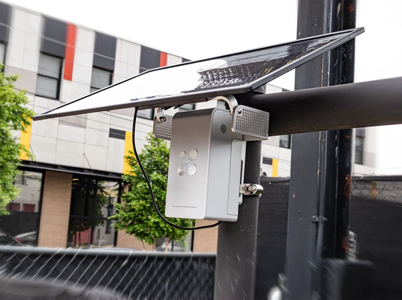 Aethair PRO under its solar panel on the Solar Panel Bracket, clamped to a street pole, an apartment building behind.