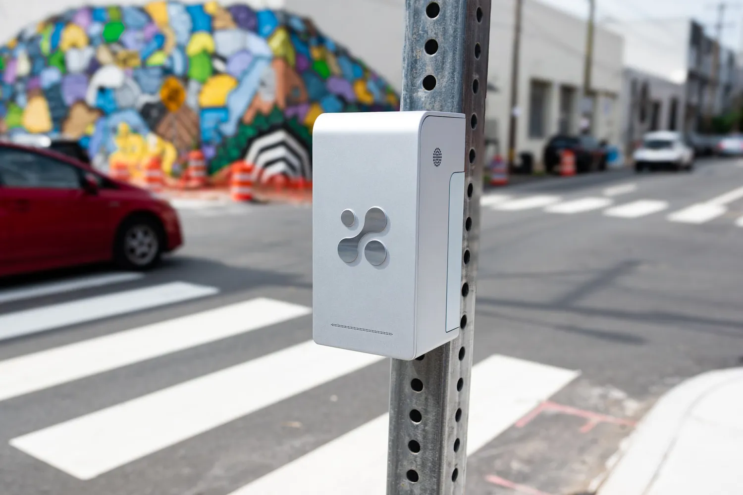Aethair PRO mounted on a street sign pole at a crosswalk, with a painted mural and parked cars behind it.