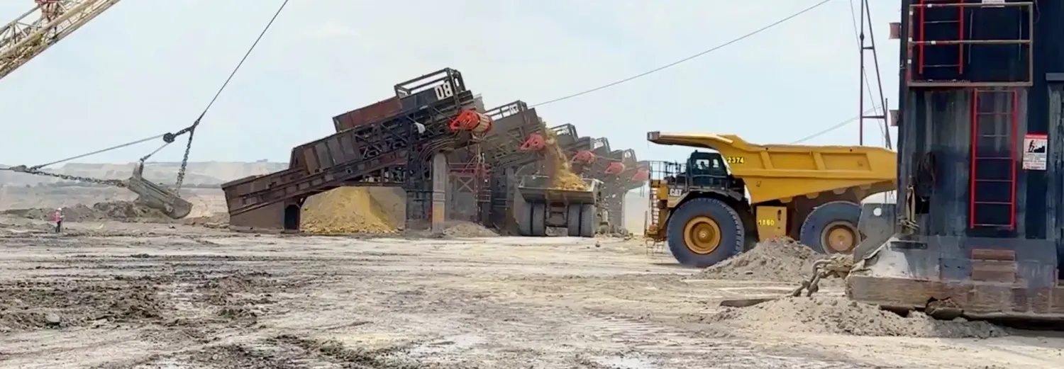 An open-pit coal mine: a dragline bucket, a row of loading hoppers, and a yellow haul truck on dusty ground.