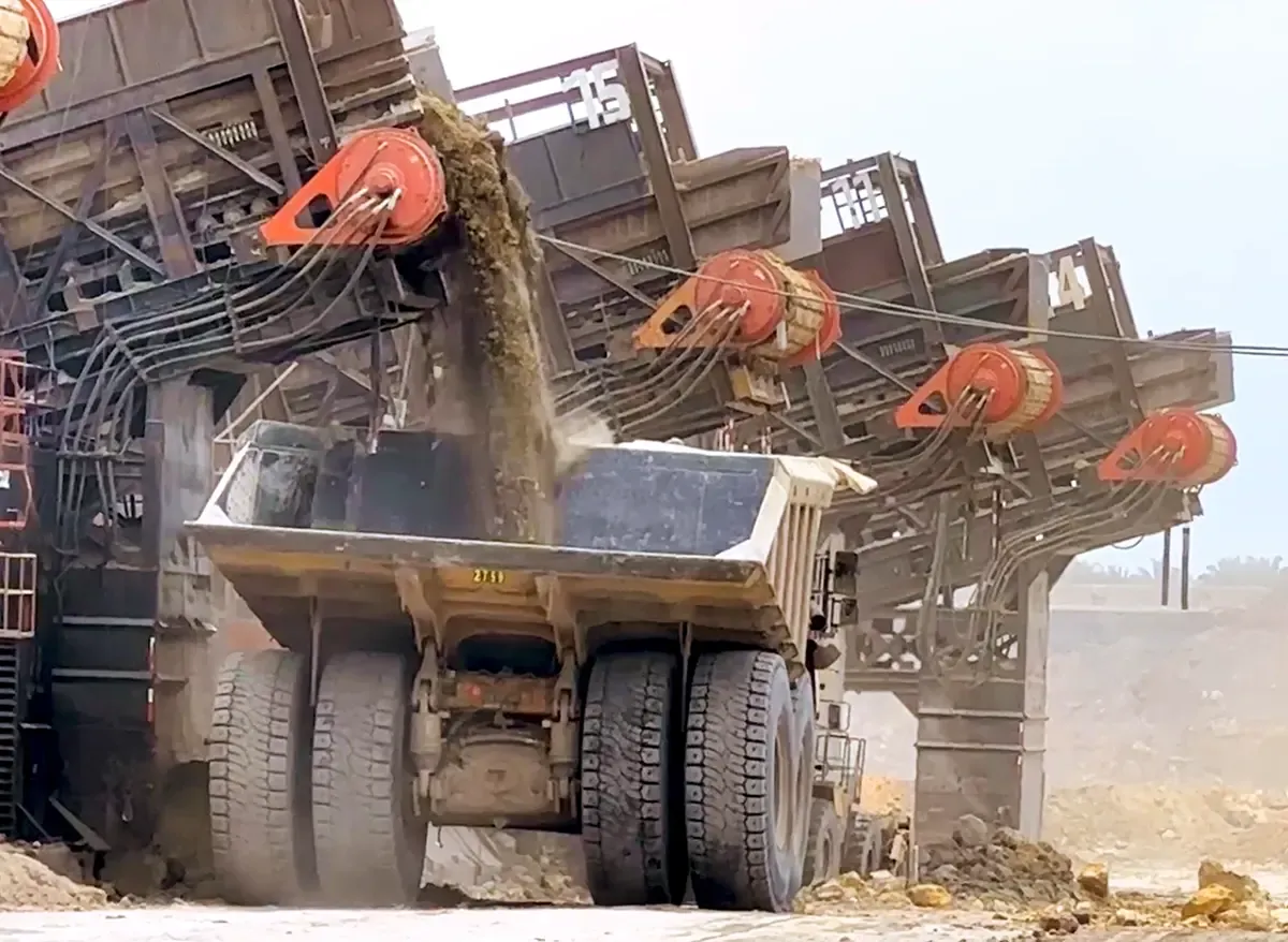 Mining equipment dumping material into a large haul truck, with dust rising around the bed.