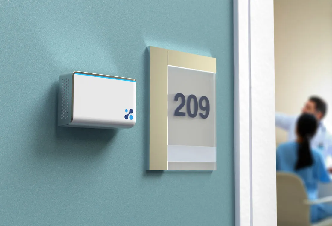 Aethair IAQ mounted next to a room number in a clinic hallway, with doctors talking in the background.