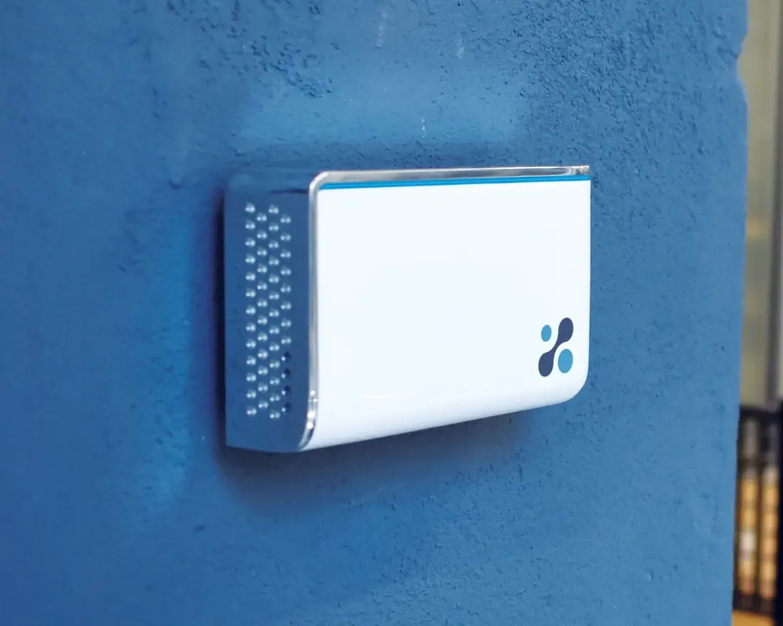 Aethair IAQ commercial indoor air quality monitor mounted on a blue wall in an open-plan office.