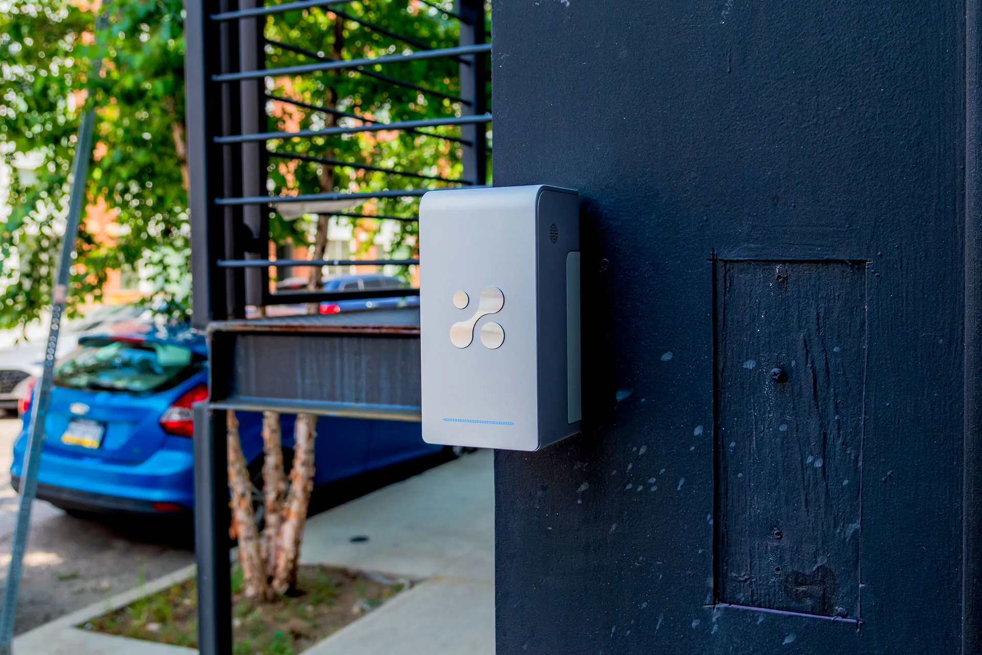 Aethair PRO mounted on a dark painted post beside a city sidewalk, with parked cars and trees behind it.